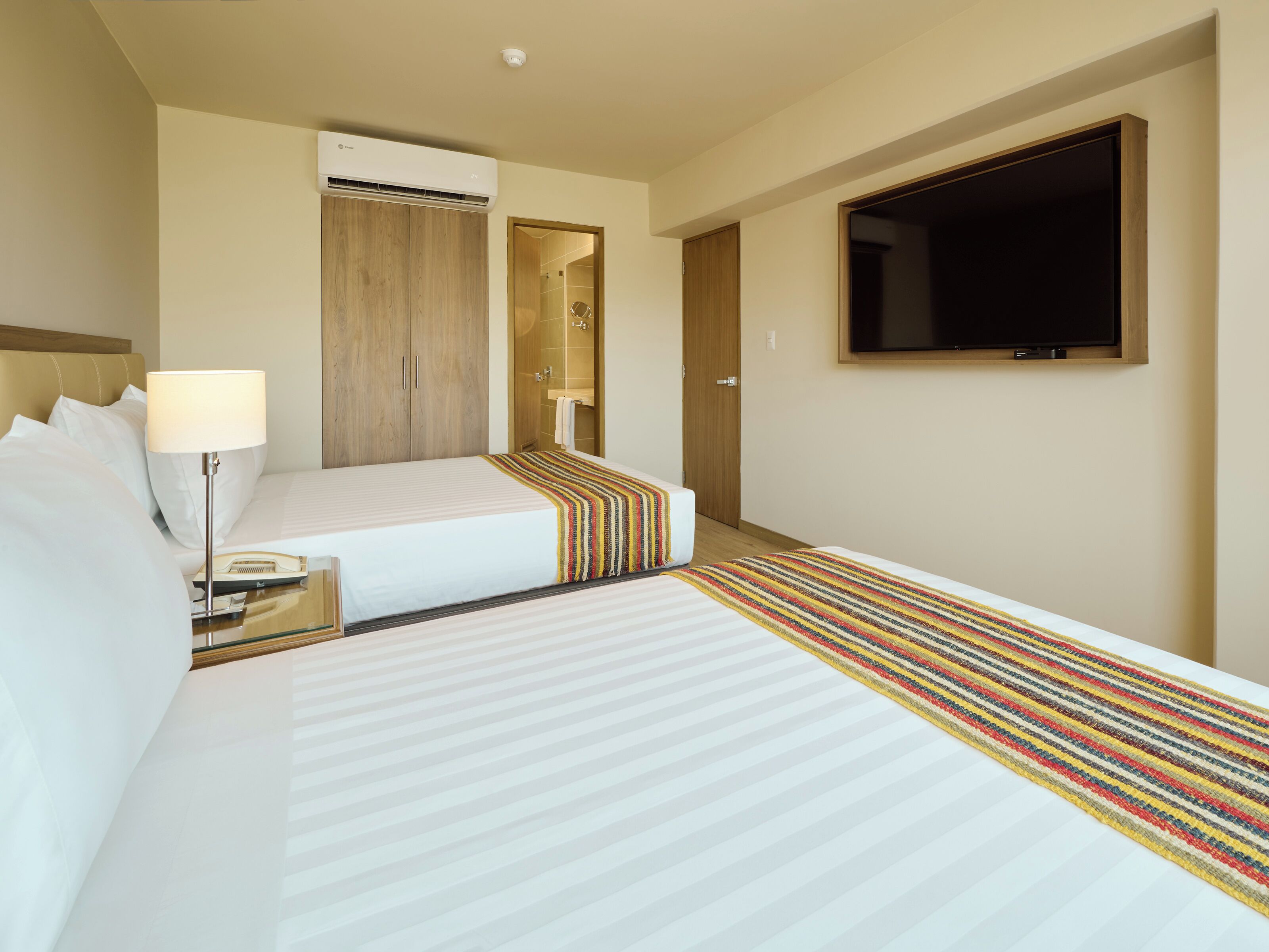 Habitación de hotel con dos camas, televisión, aire acondicionado, lámpara de noche y baño visible a través de una puerta abierta.