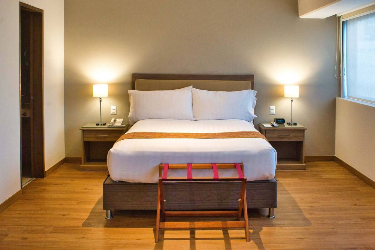 Een hotelkamer met een groot bed, nachtkastjes met lampen, een houten bankje en een raam met zonwering.