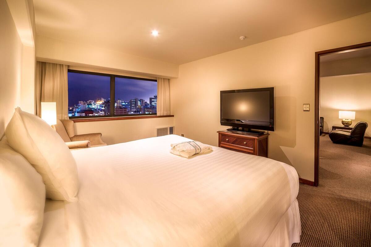 Hotelkamer met een groot bed, een televisie, een bureau met lamp en uitzicht op de stad 's nachts.
