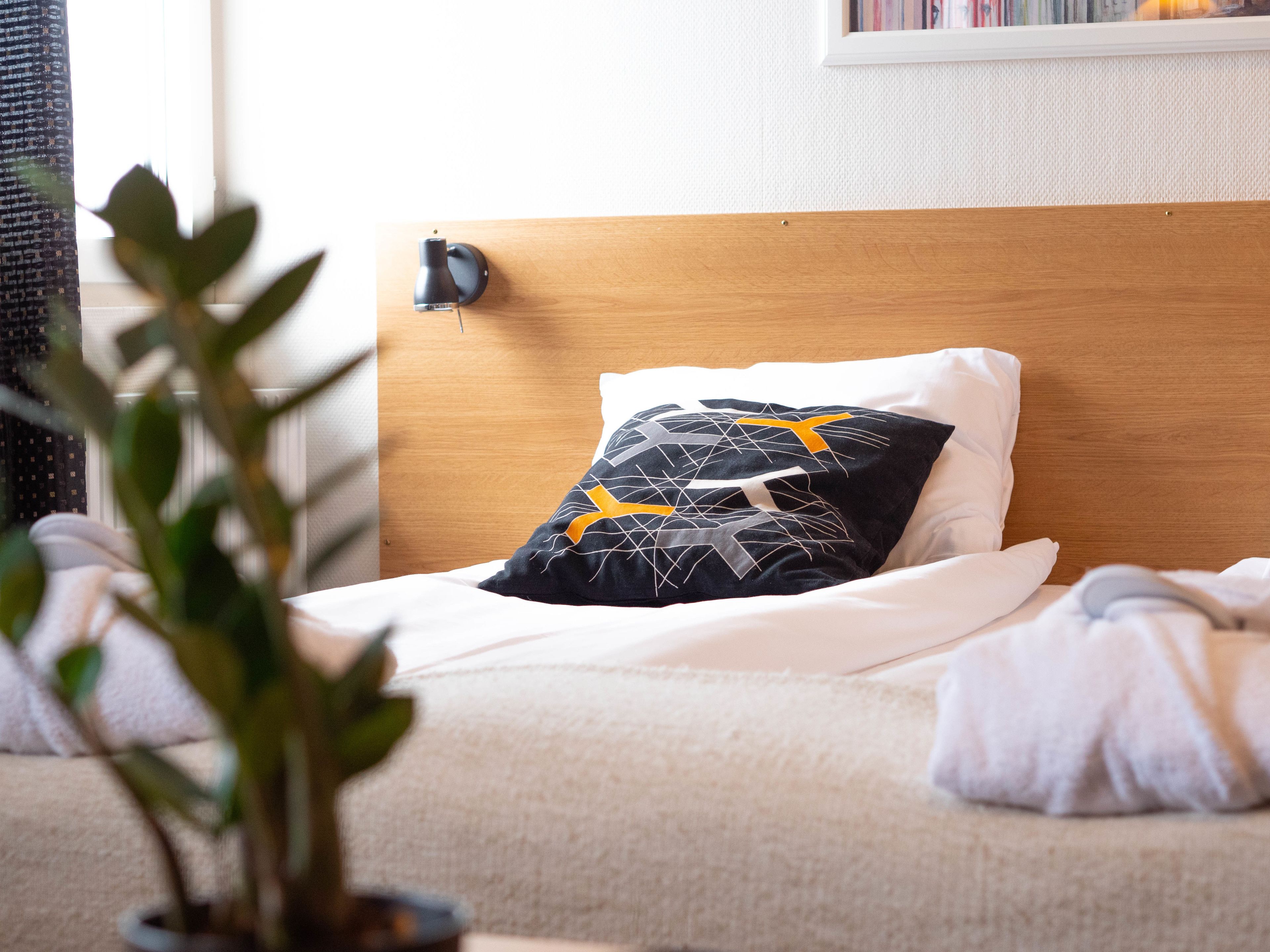 Een net opgemaakt bed met een houten hoofdeinde, een decoratief kussen en een bedlampje.