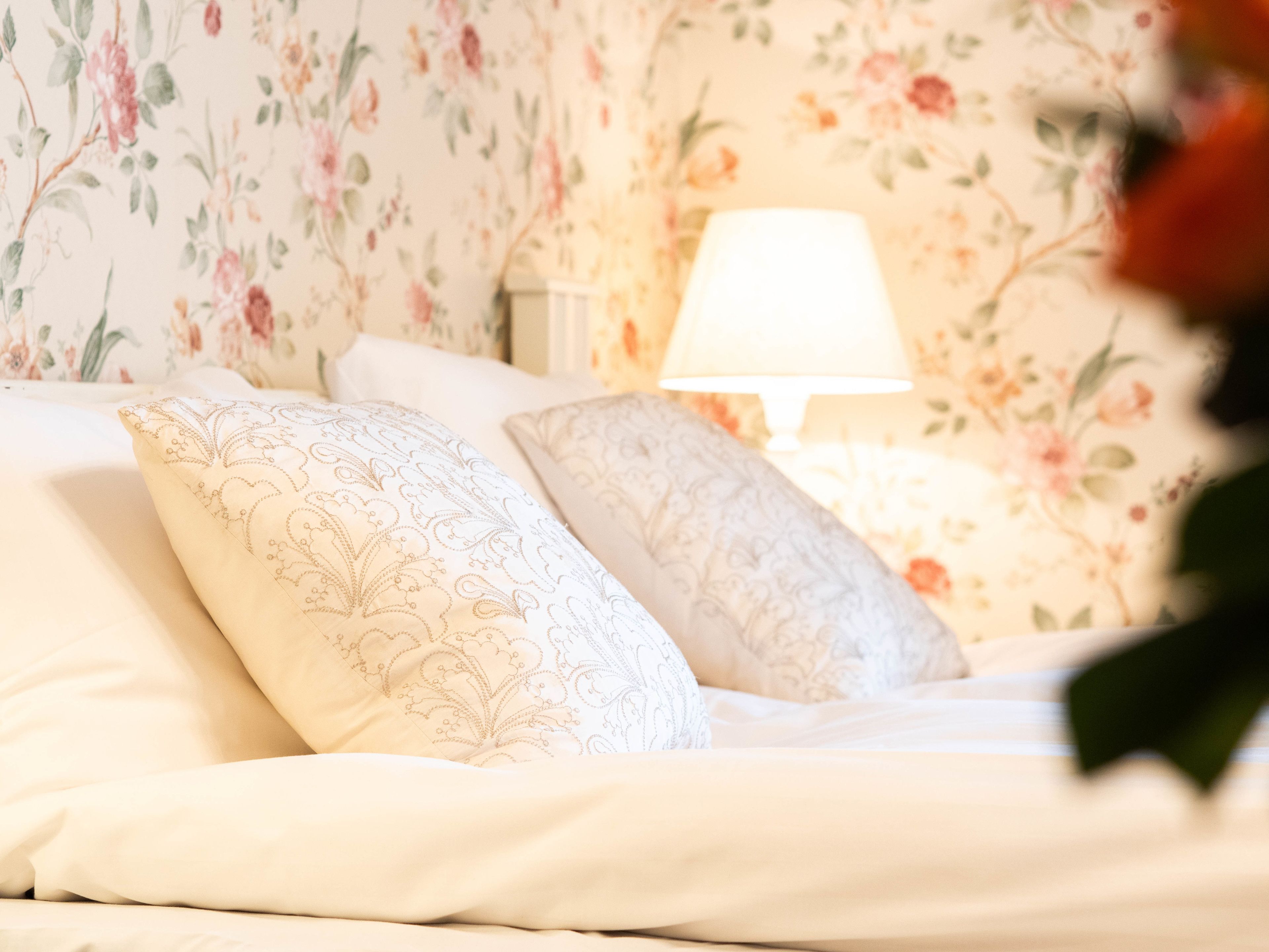 En seng med blomstret tapet, to mønstrede puder og en sengetabletlampe.