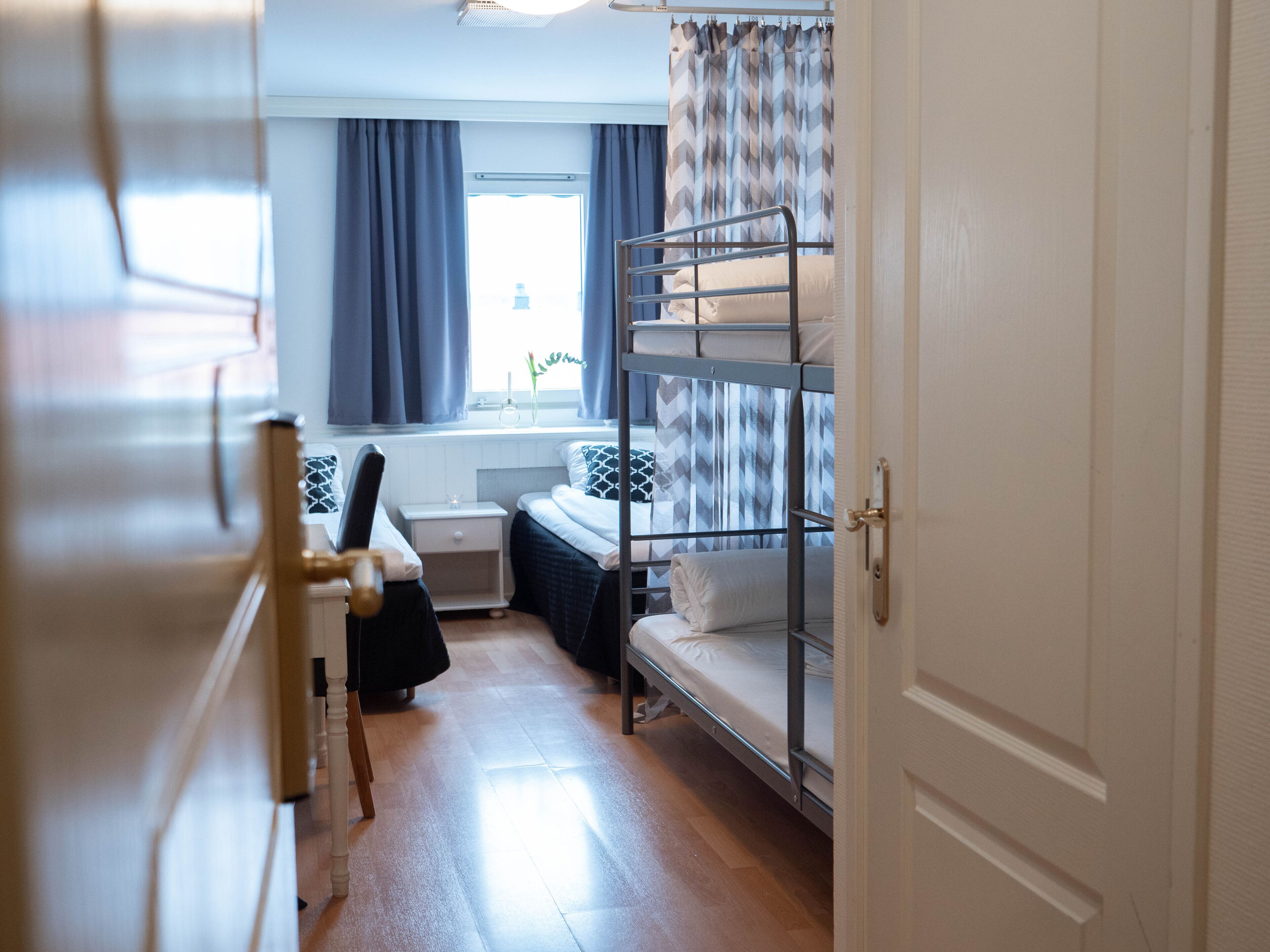 Habitación de hotel pequeña con una litera, una mesita de noche, una silla y un ventanal con cortinas.