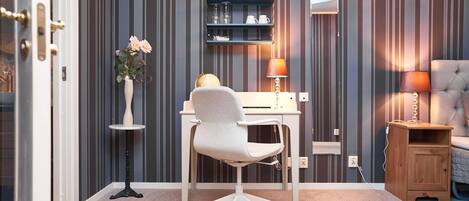 A room with a white desk, a white office chair, a small wooden nightstand, a lamp, a vase with flowers, and a shelf with books and decorative items.