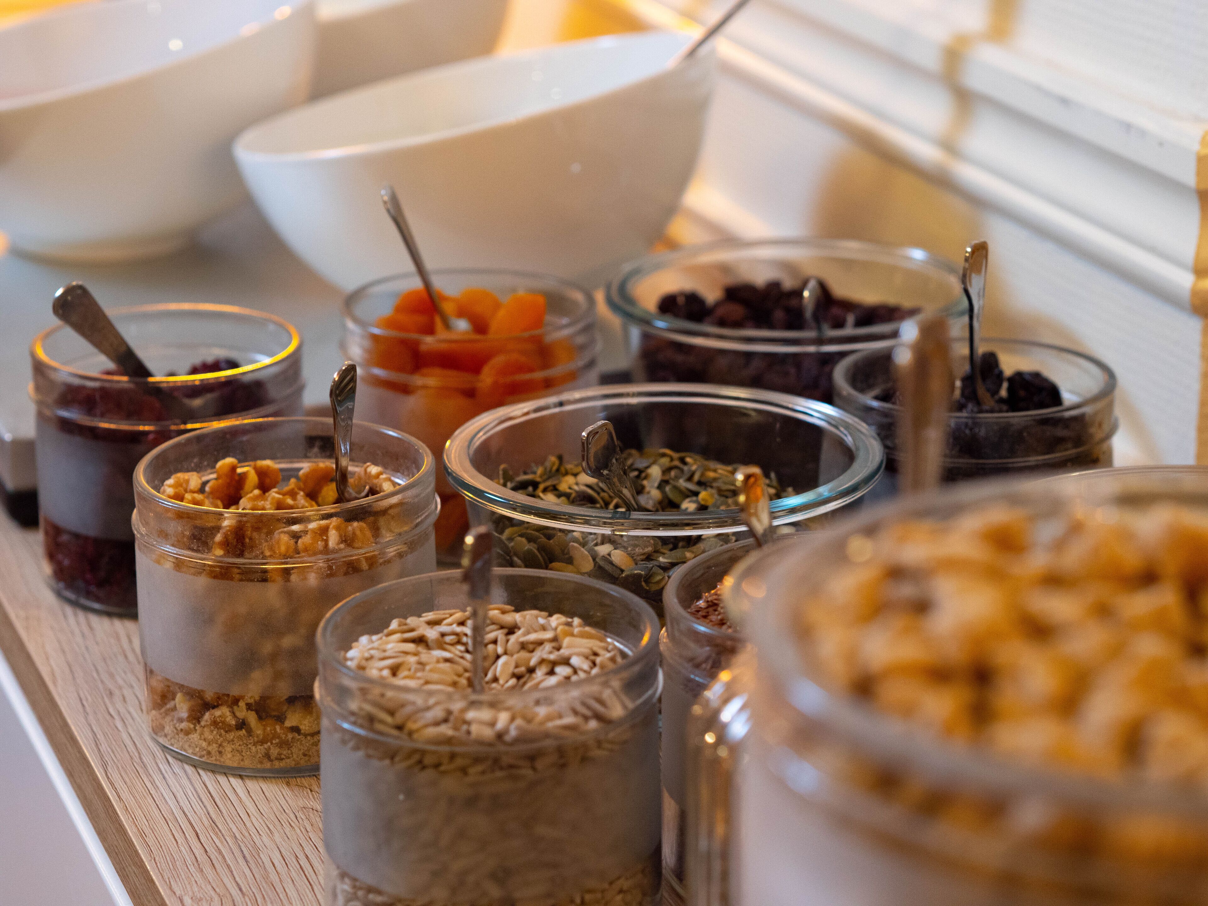 En buffet med forskellige madvarer i glasbeholdere, herunder nødder, frø og tørret frugt.
