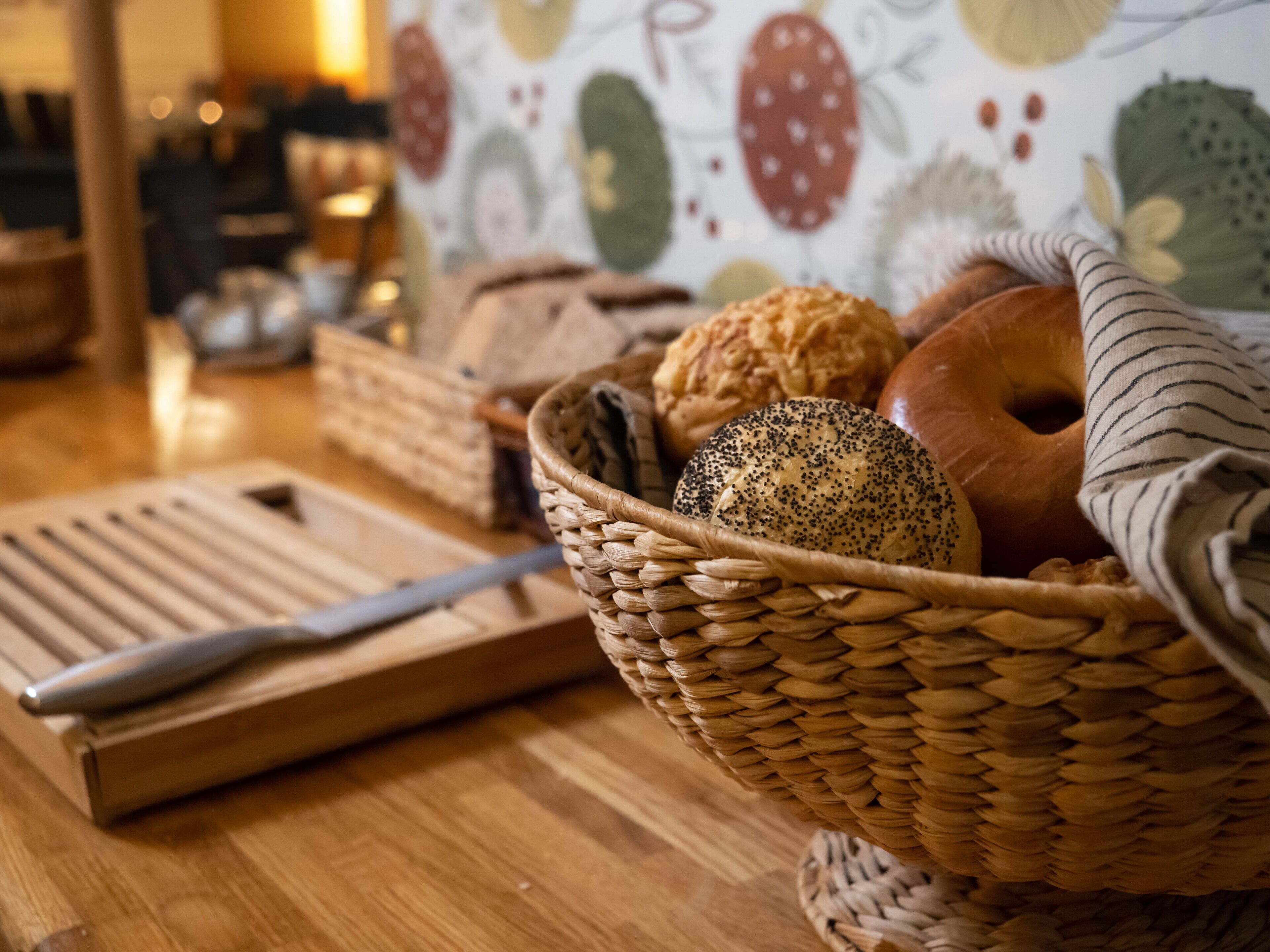 A basket of various breads and a cutting board with a knife on a wooden table.