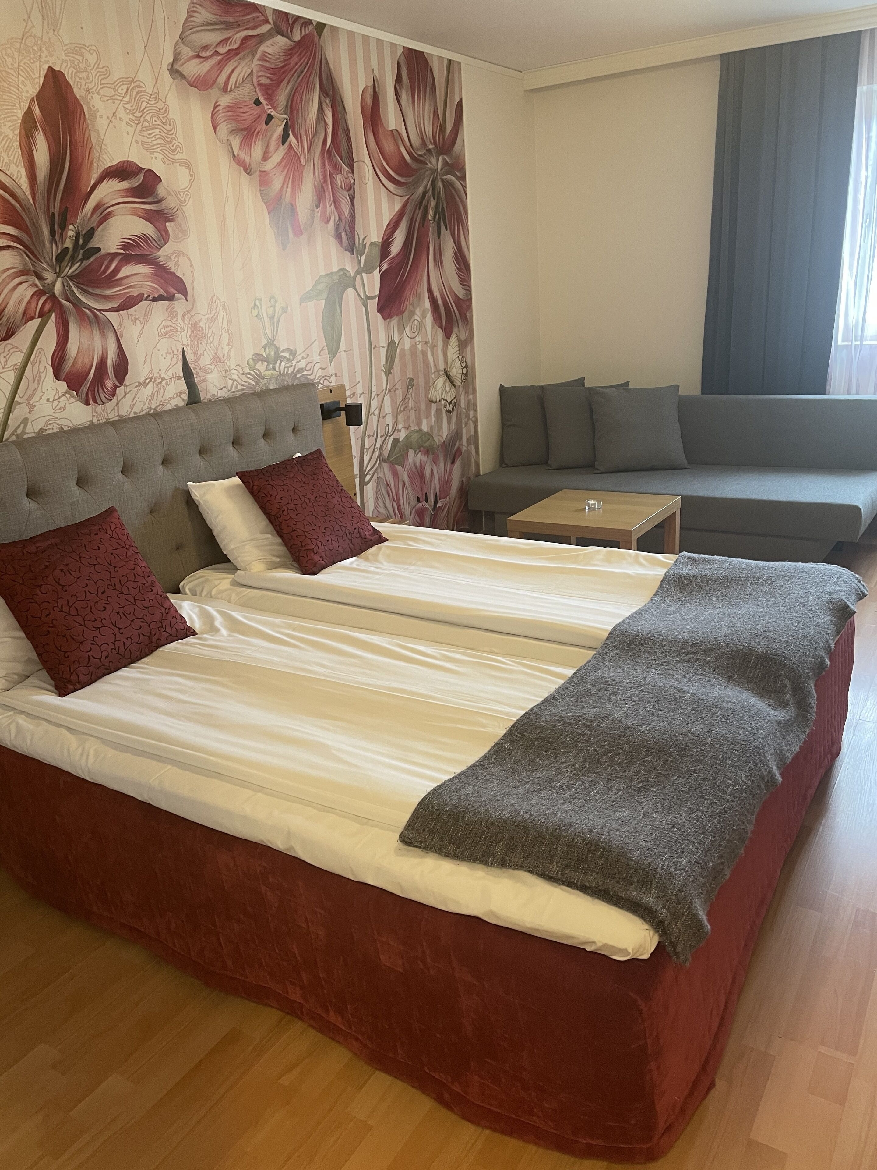 A double bed with a grey blanket, two red pillows, and a floral wallpaper.