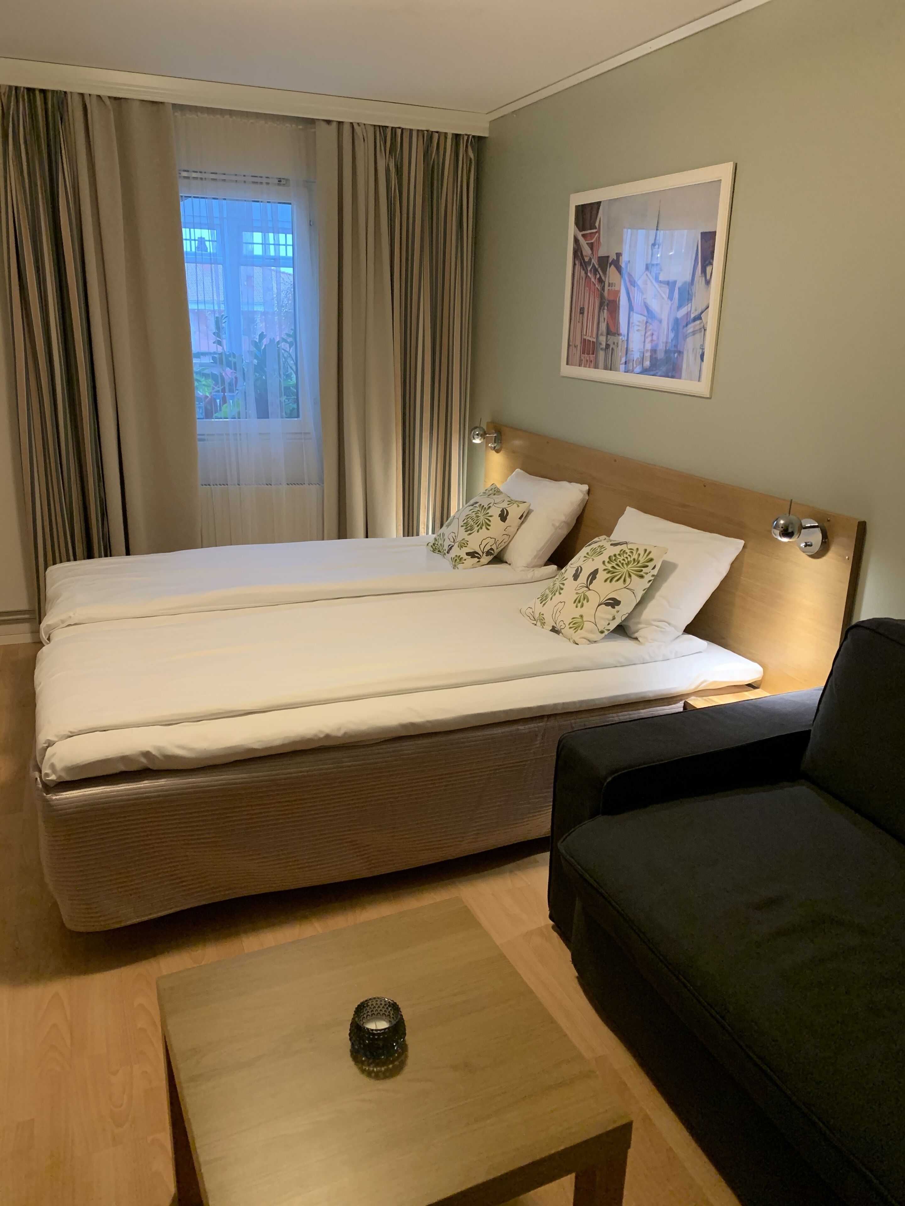 A hotel room with a double bed, a bedside table with a lamp, a dark-colored sofa, and a window with curtains.
