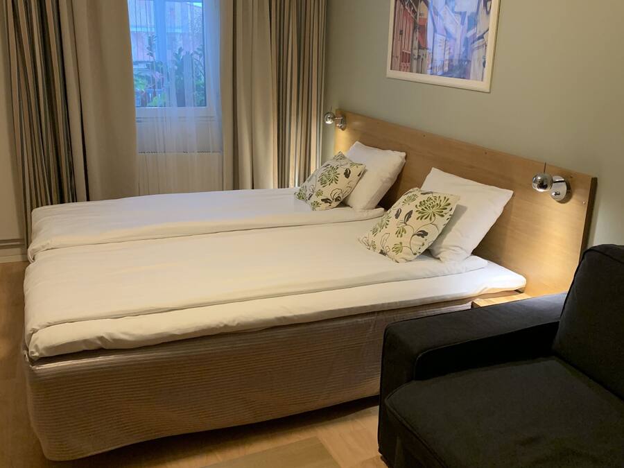 A hotel room with a double bed, a bedside table with a lamp, a dark-colored sofa, and a window with curtains.