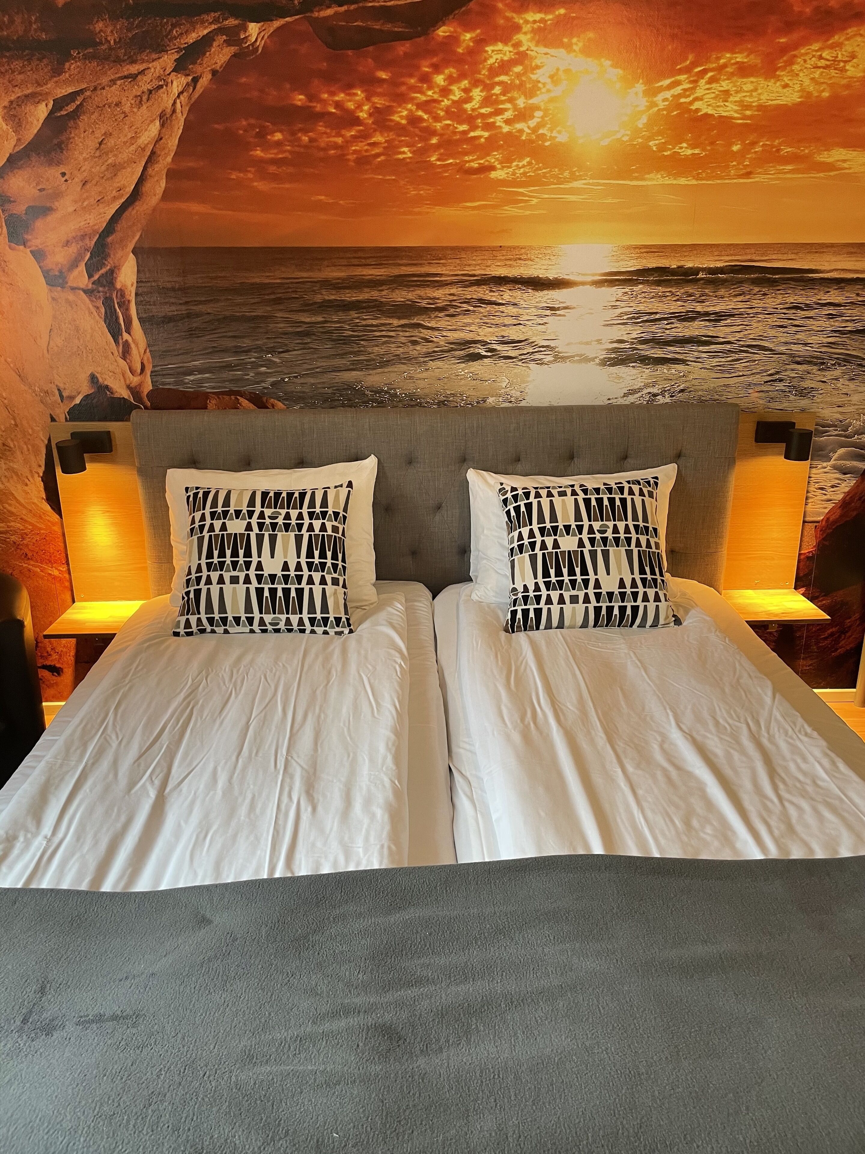 A double bed with white linens and patterned pillows, set against a backdrop of a sunset over the ocean, framed by a rock formation.