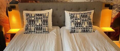A double bed with white linens and patterned pillows, set against a backdrop of a sunset over the ocean, framed by a rock formation.