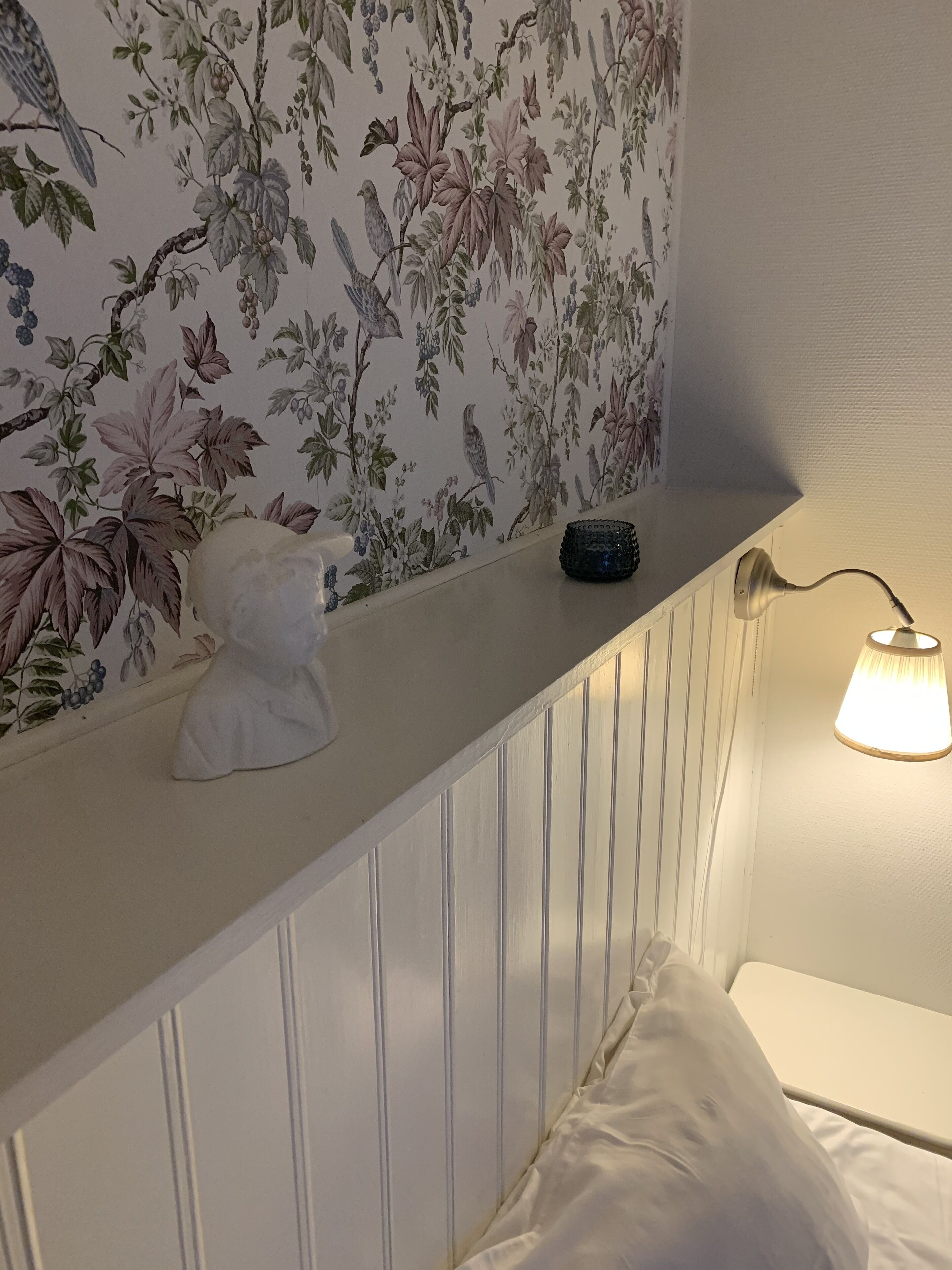 Un baño con papel tapiz floral, un tocador blanco, una pequeña escultura de busto y una lámpara de noche.