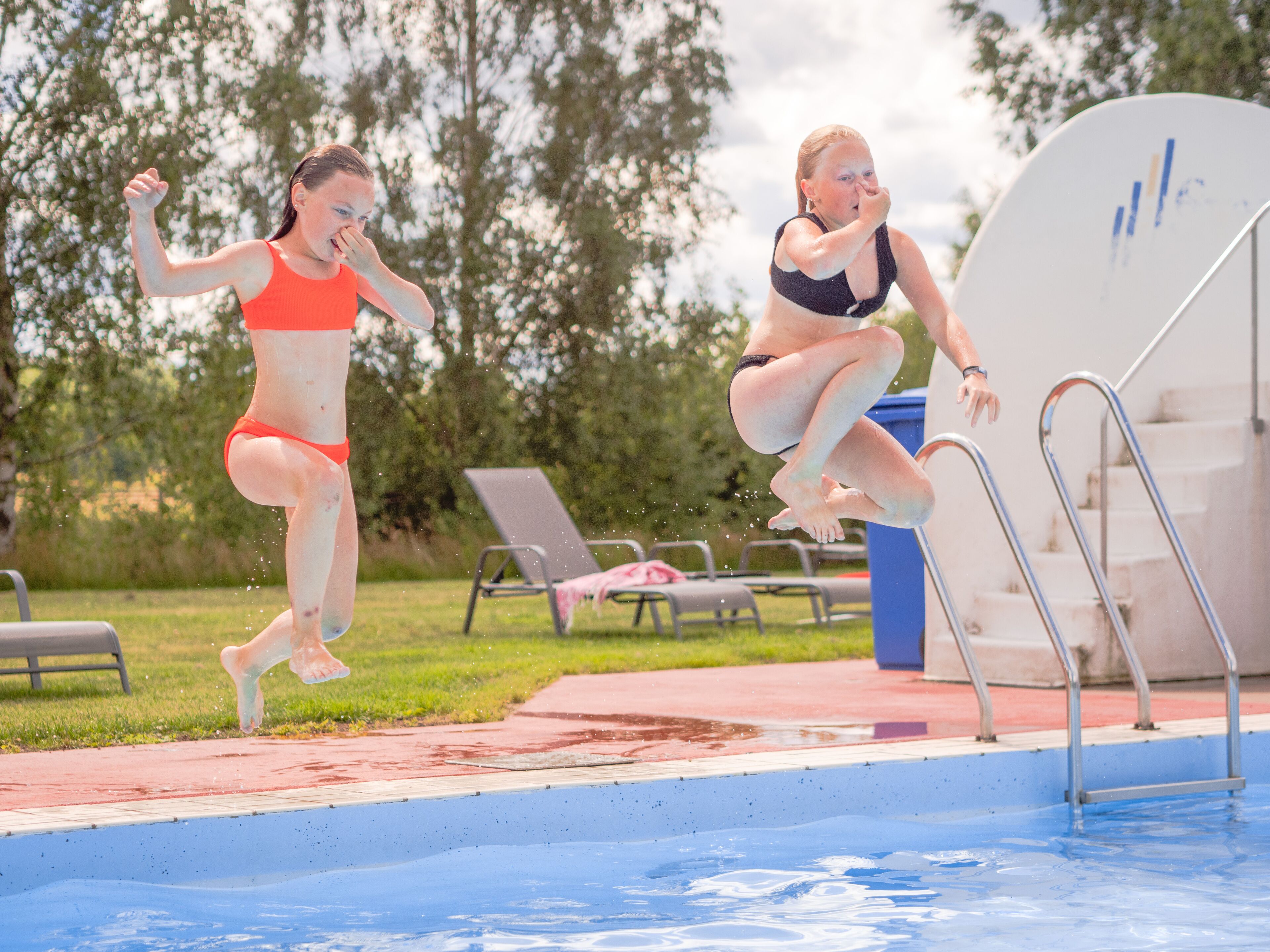 Deux personnes sautent dans une piscine, avec un plongeoir et des chaises longues en arrière-plan.