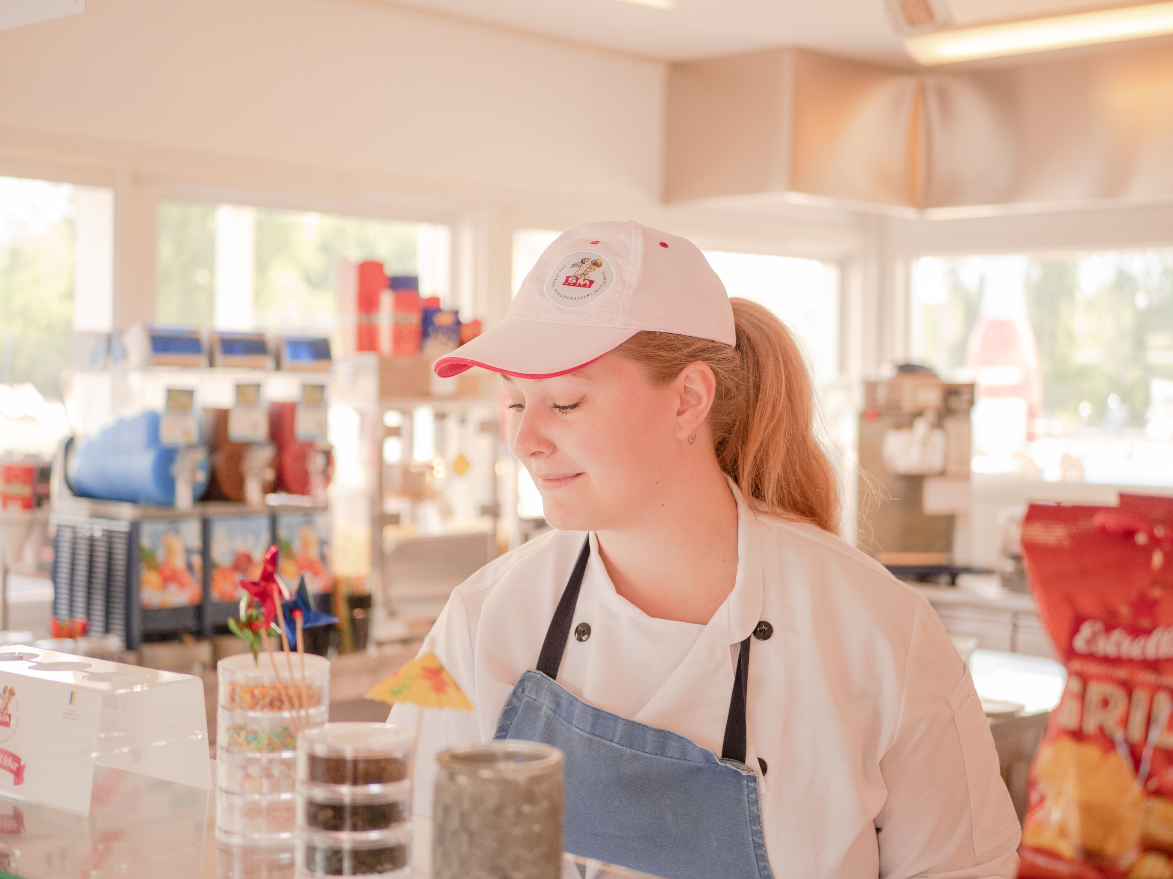 A person wearing a white shirt and a cap, working at a counter with various items like jars and a bag of chips.
