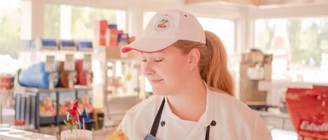 A person wearing a white shirt and a cap, working at a counter with various items like jars and a bag of chips.