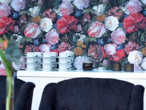 Papel tapiz floral con una pila de tazas y platillos blancos, una jarra de café y dos tazas estampadas sobre una mesa.
