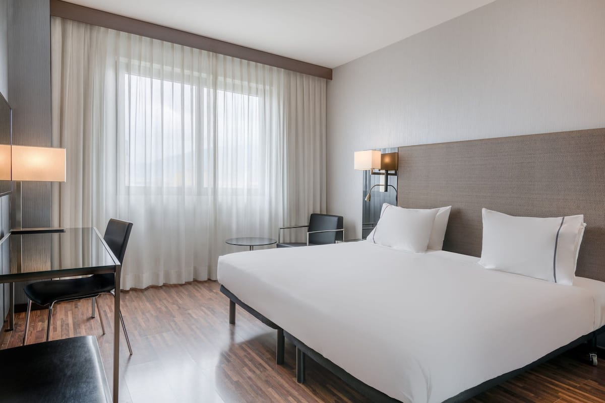 Una habitación de hotel con una cama grande, un escritorio con una silla, una mesita y una lámpara.