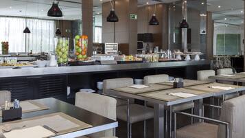 Uma área de refeições moderna com mesas, cadeiras, um buffet com comida e bebidas, e um espelho grande que reflete o espaço.
