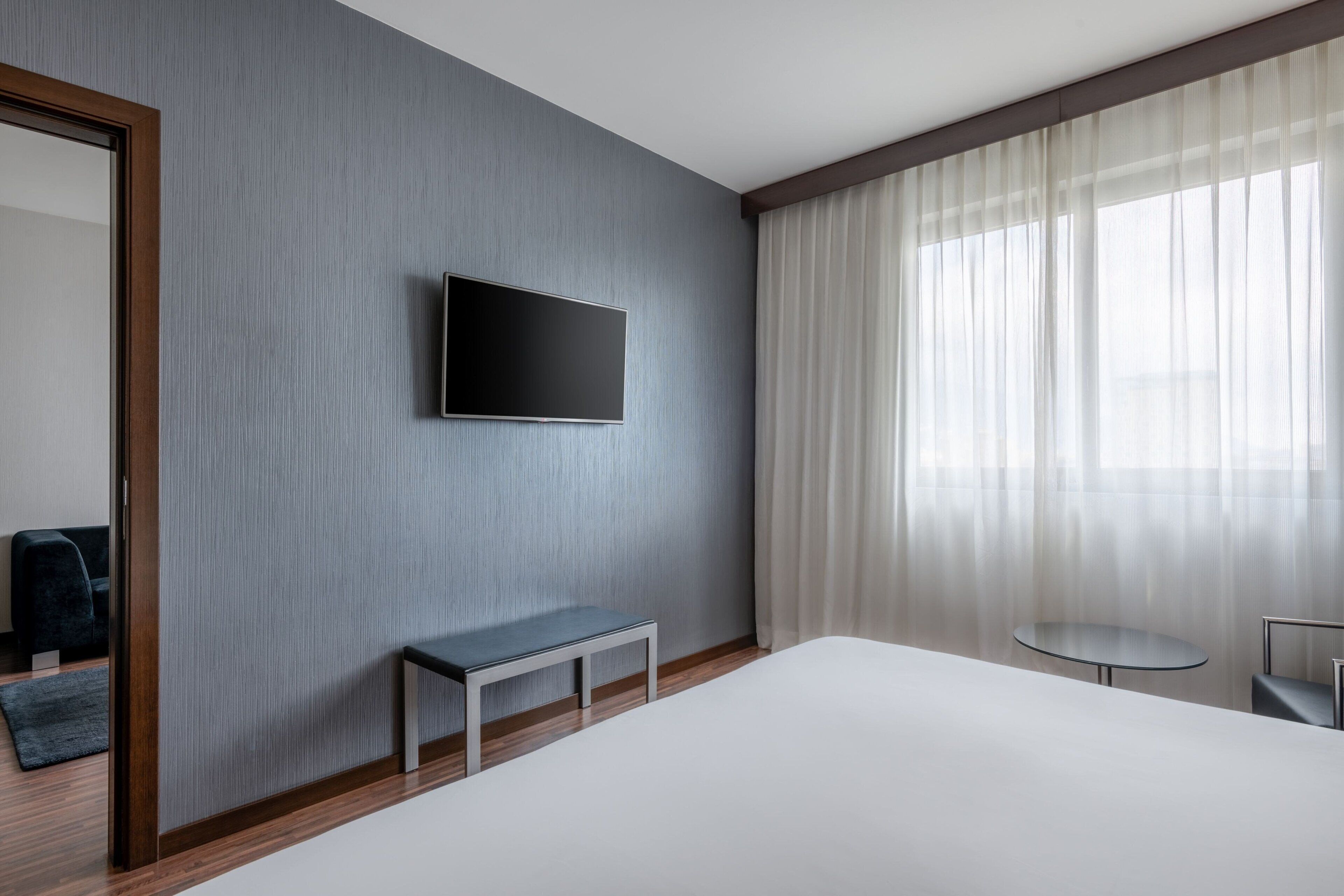 A modern hotel room with a large bed, a small table, a bench, a flat-screen TV mounted on the wall, and a large window with sheer curtains.