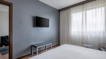 A modern hotel room with a large bed, a small table, a bench, a flat-screen TV mounted on the wall, and a large window with sheer curtains.