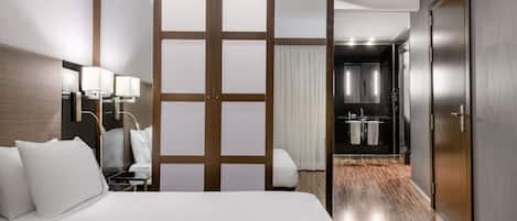 A hotel room with a large bed, a desk with a lamp, a sliding door leading to a bathroom, and a wooden floor.