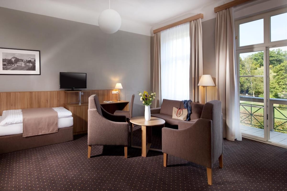Ein Hotelzimmer mit Bett, einer Couch, einem kleinen Tisch mit Vase, einem Fernseher und einem Fenster mit Vorhängen.
