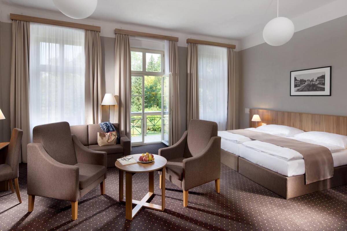 Ein Hotelzimmer mit zwei Sesseln, einem kleinen Tisch mit Obstschale, einem Bett mit weißer Bettwäsche und einem großen Fenster mit Vorhängen.