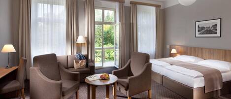 A hotel room with two armchairs, a small table with a bowl of fruit, a bed with white linens, and a large window with curtains.