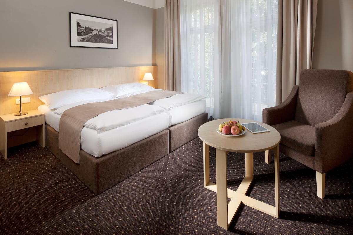 Ein Hotelzimmer mit Bett, Nachttischen, einem Sessel, einem kleinen Tisch mit Früchten und einem Tablet, sowie einem gerahmten Bild an der Wand.