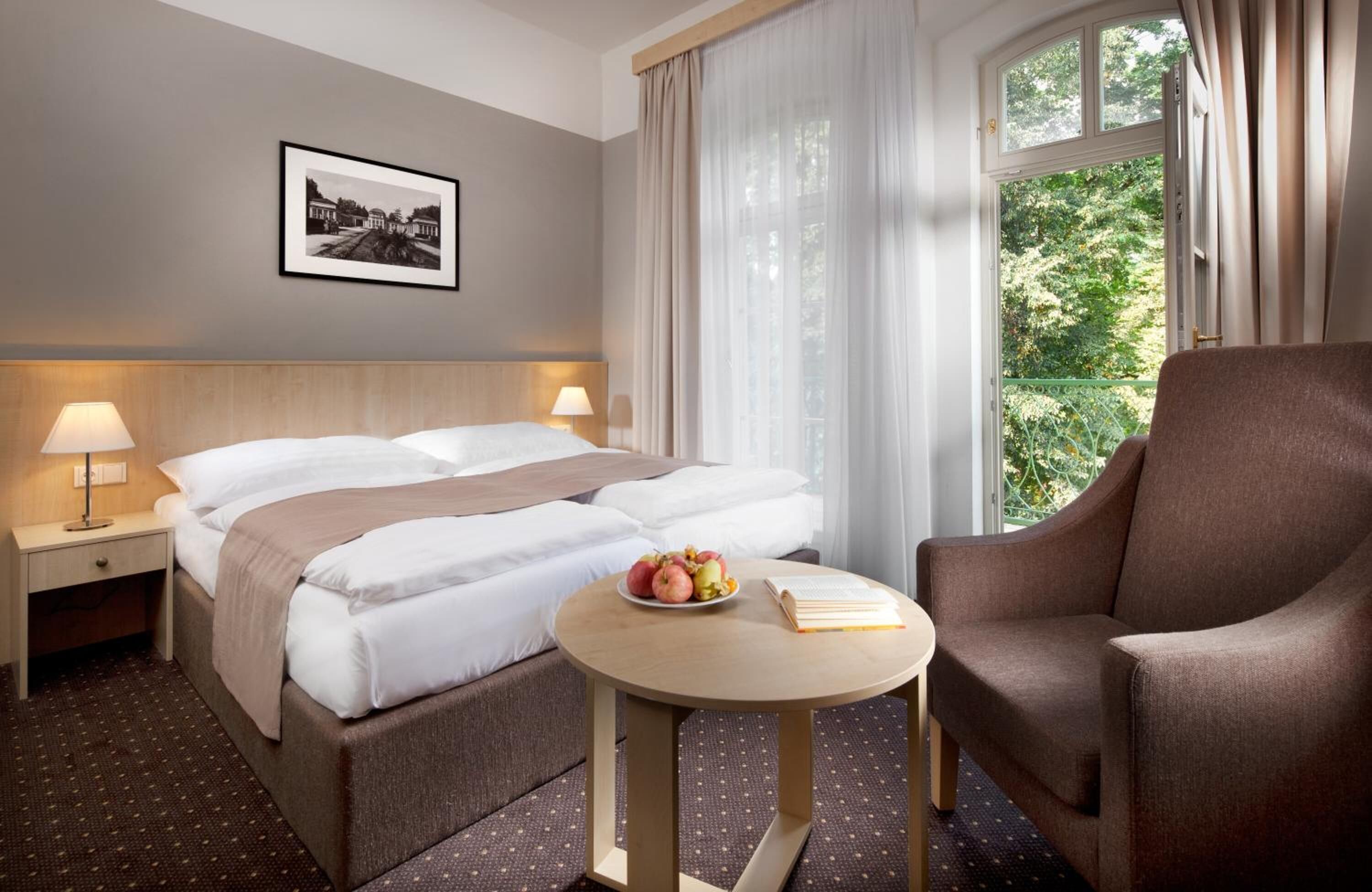 A hotel room with a bed, bedside lamps, a round table with a book and fruit, a chair, and a window with curtains.