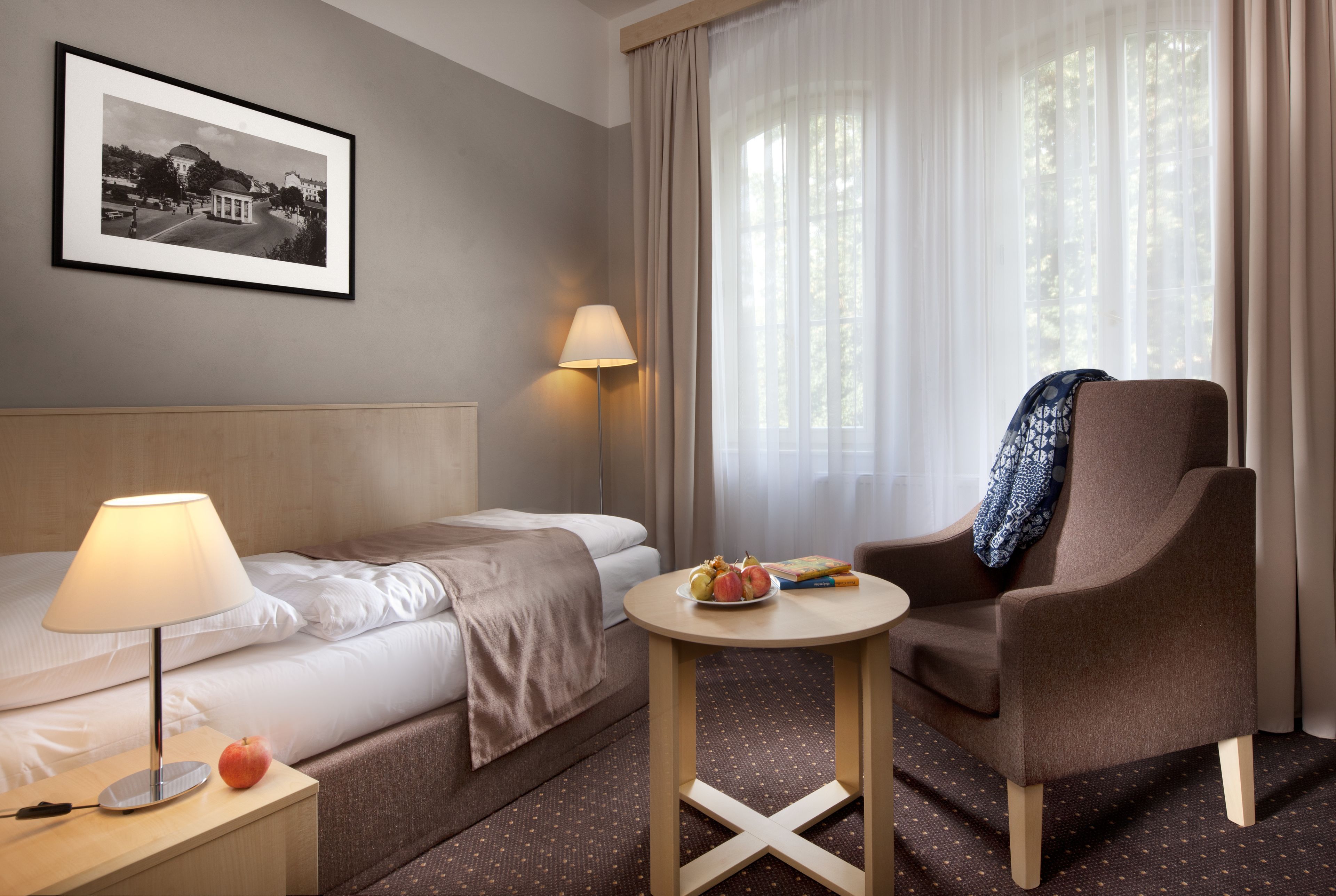 Una habitación de hotel con una cama, una silla, una mesita con frutas, una lámpara y un cuadro en la pared.