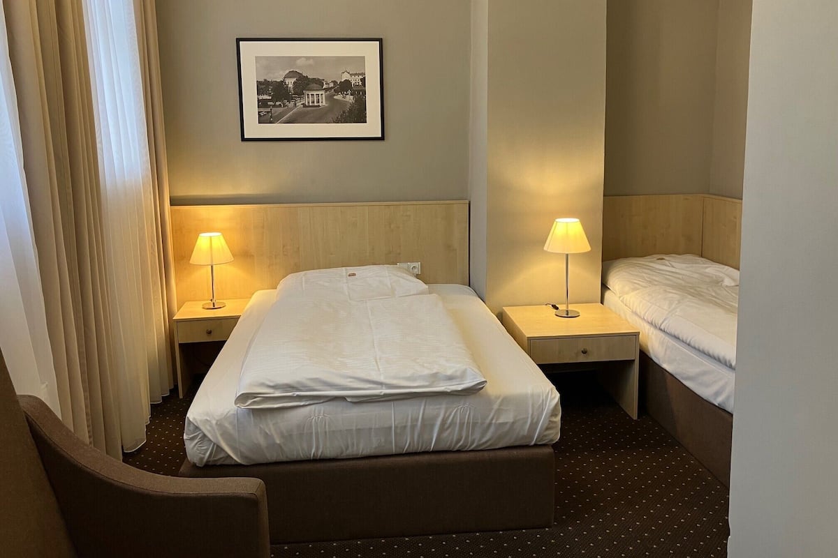 Ein Hotelzimmer mit einem Bett, zwei Nachttischen mit Lampen, einem Kopfteil, einem gerahmten Bild an der Wand und einem Fenster mit Vorhängen.