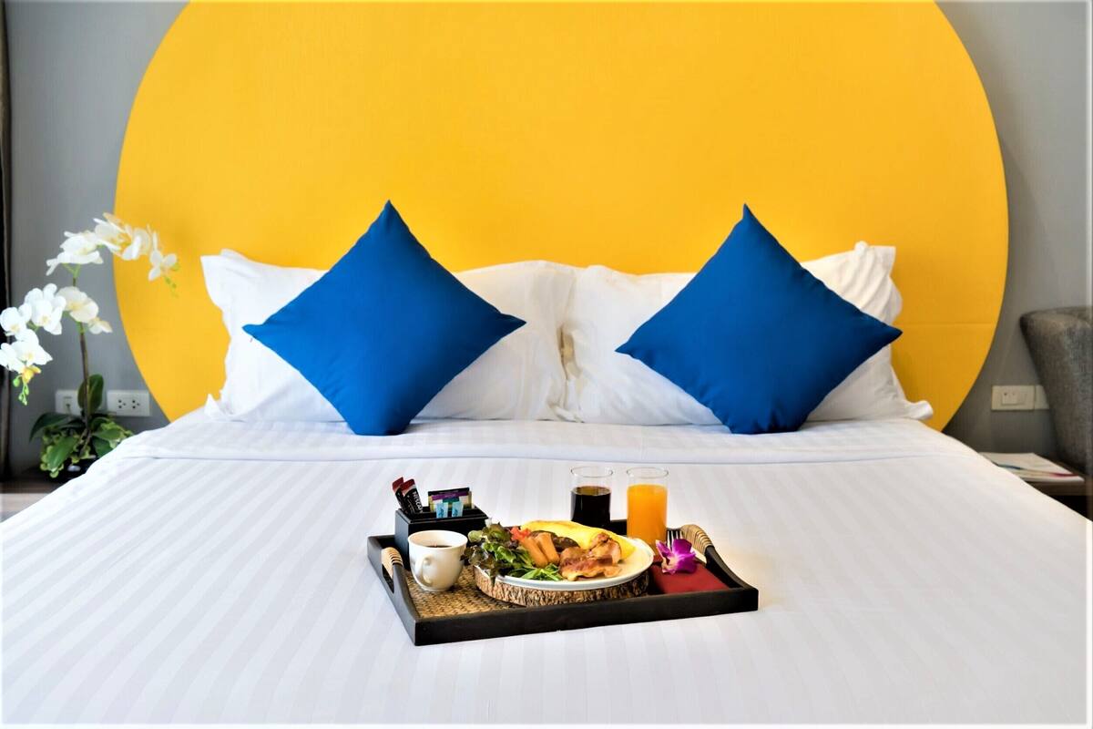 A neatly made bed with a tray of breakfast items, including eggs, toast, and juice, in front of a large yellow wall with blue pillows.