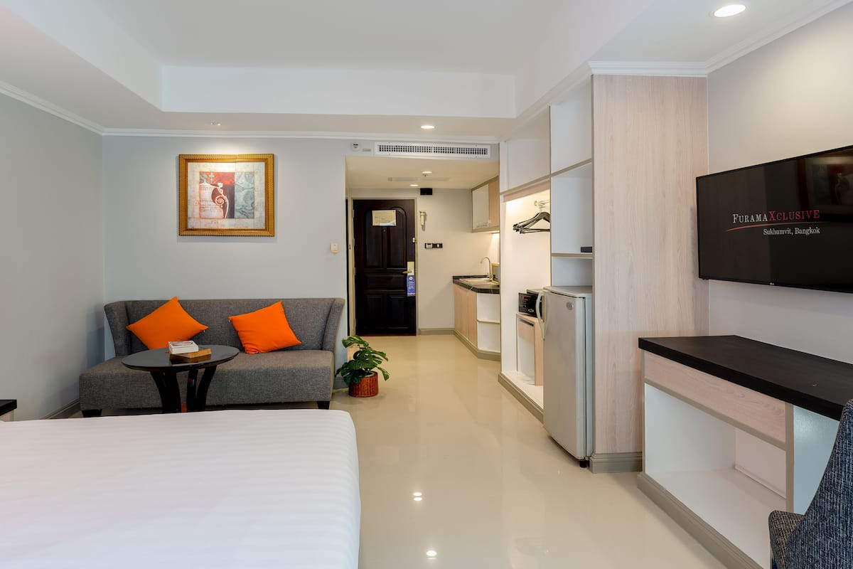 A hotel room with a bed, a sofa with orange cushions, a small table, a TV, a kitchenette, and a bathroom.