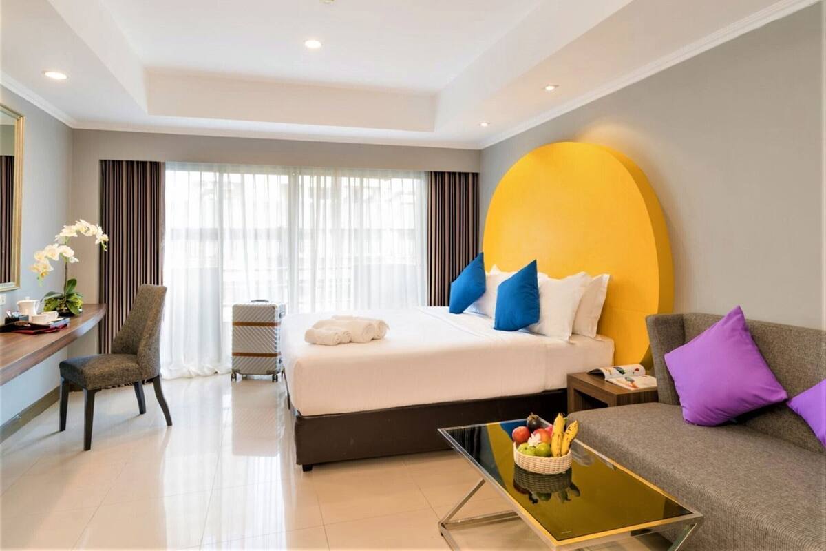 A hotel room with a bed, a sofa, a glass coffee table, a desk with a chair, and a window with curtains.