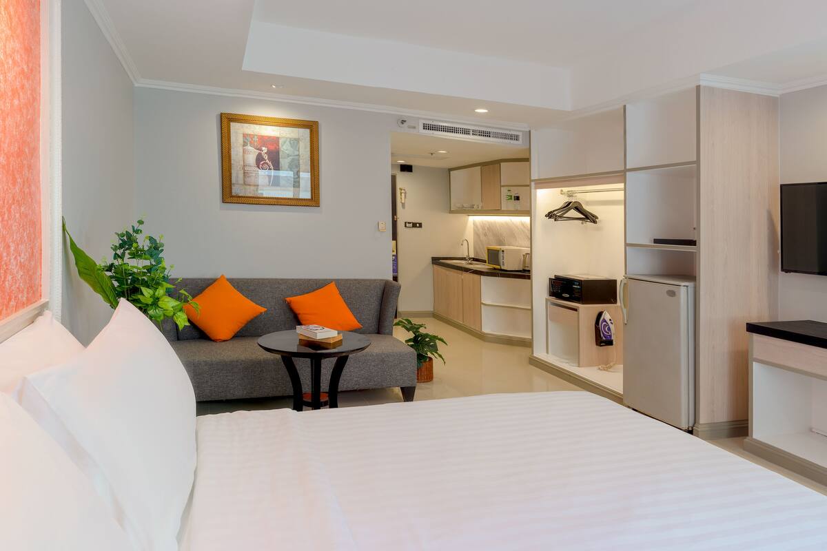 A hotel room with a bed, a sofa with orange cushions, a small table with a book, a plant, and a kitchen area with a microwave and refrigerator.