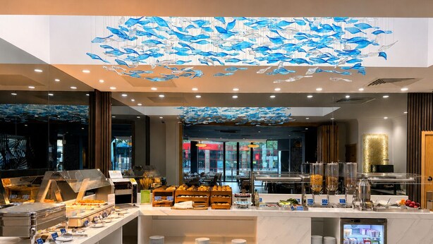 A modern hotel lobby with a central buffet area, a refrigerator, and a large ceiling mural of fish.