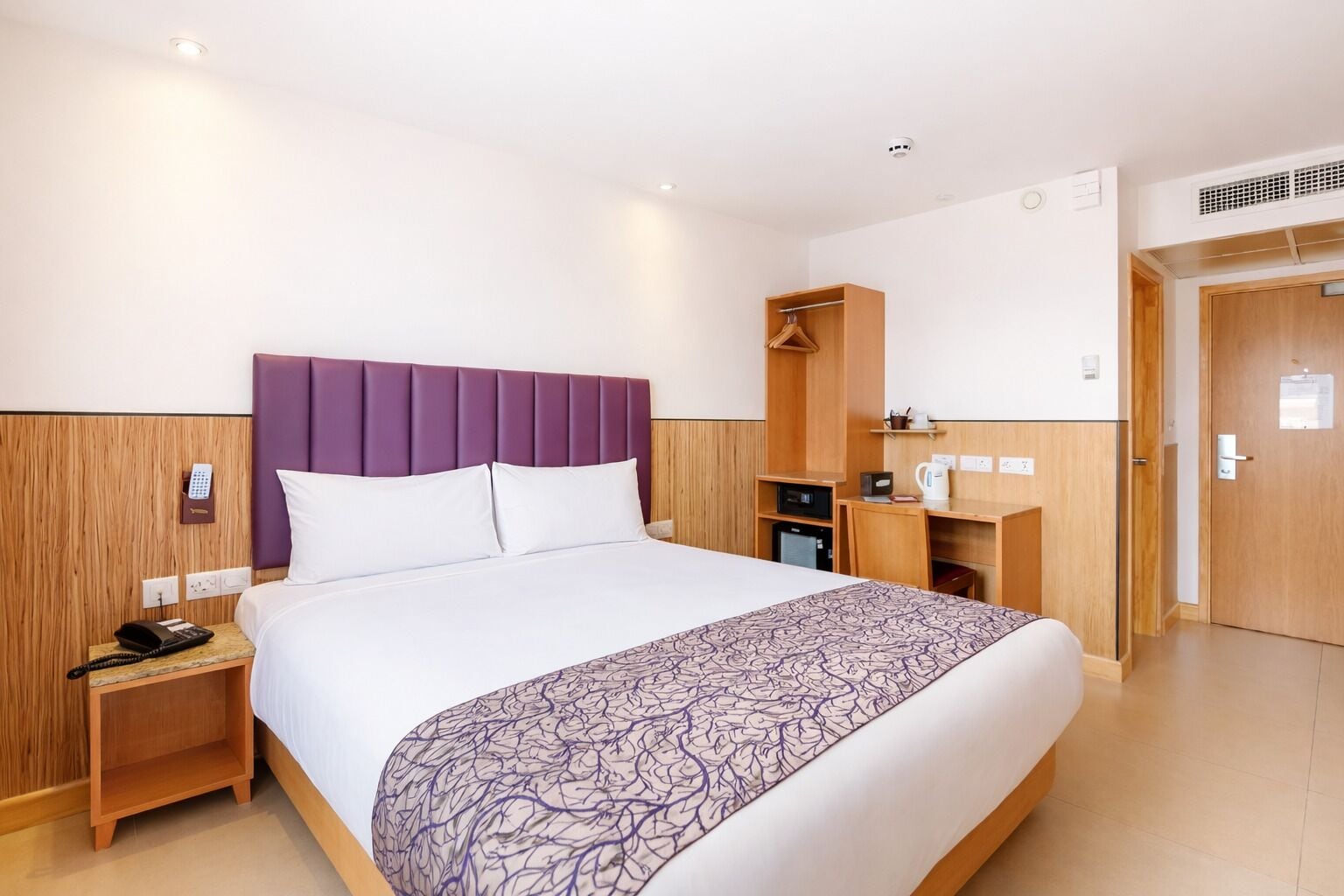 A hotel room with a bed, a wooden nightstand, a wardrobe, a desk with a chair, and a door.