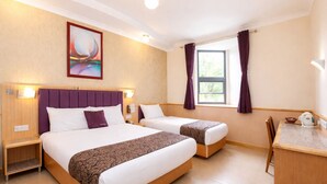 A hotel room with two beds, a desk, a chair, a window with curtains, a painting on the wall, and a nightstand with a lamp.