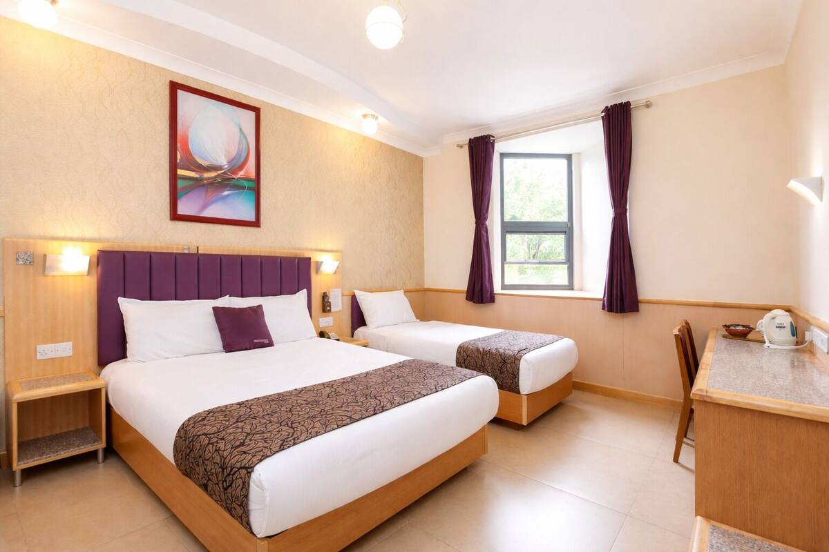 Ein Hotelzimmer mit zwei Betten, einem Schreibtisch, einem Stuhl, einem Fenster mit Vorhängen, einem Gemälde an der Wand und einem Nachttisch mit Lampe.