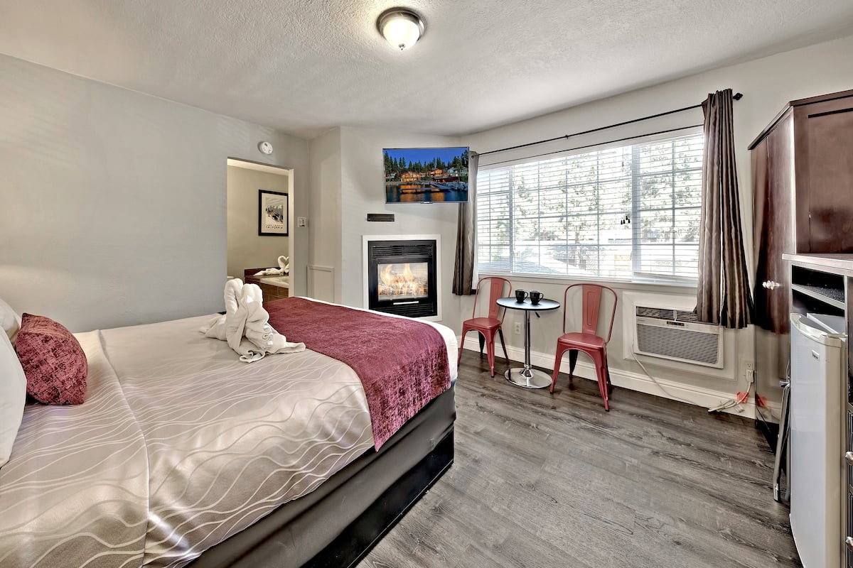 A hotel room with a bed, a fireplace, a TV, a window with curtains, and a desk with chairs.