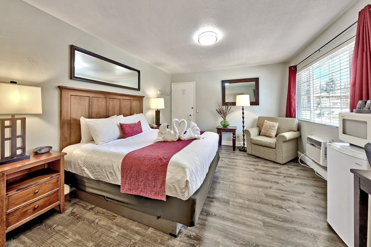 A hotel room with a bed, bedside tables, a chair, a desk, a television, and a window with blinds.