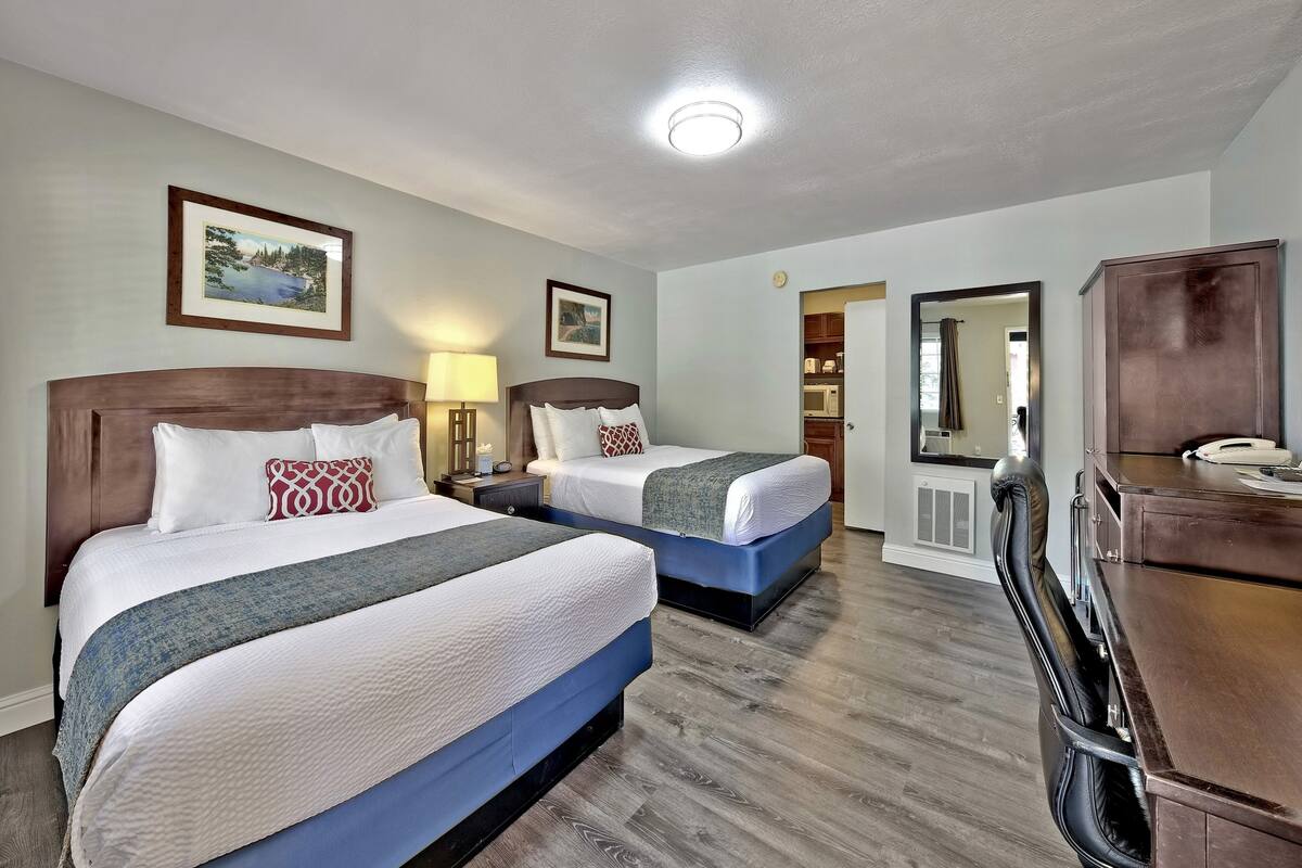 A hotel room with two beds, a desk, a chair, a mirror, and a small kitchenette.
