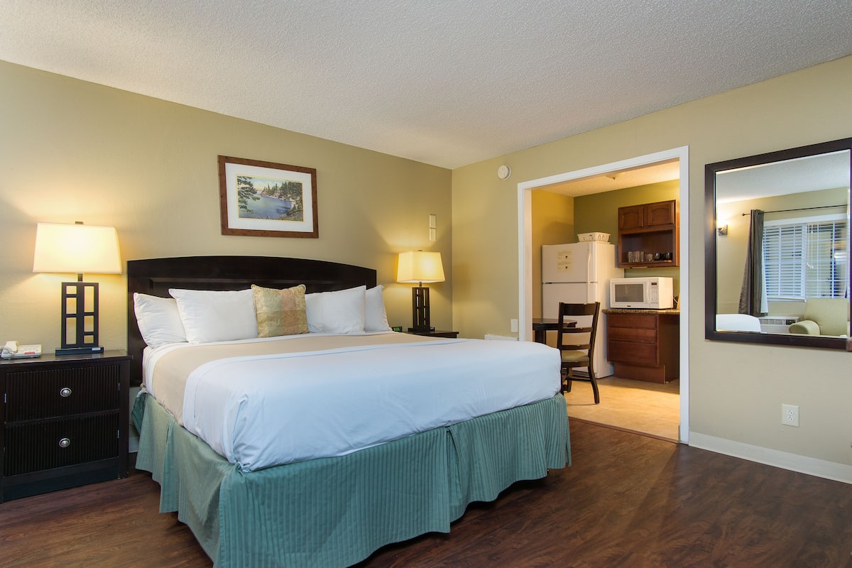 A hotel room with a large bed, two bedside lamps, a framed picture on the wall, and a view of the kitchen through an open doorway.