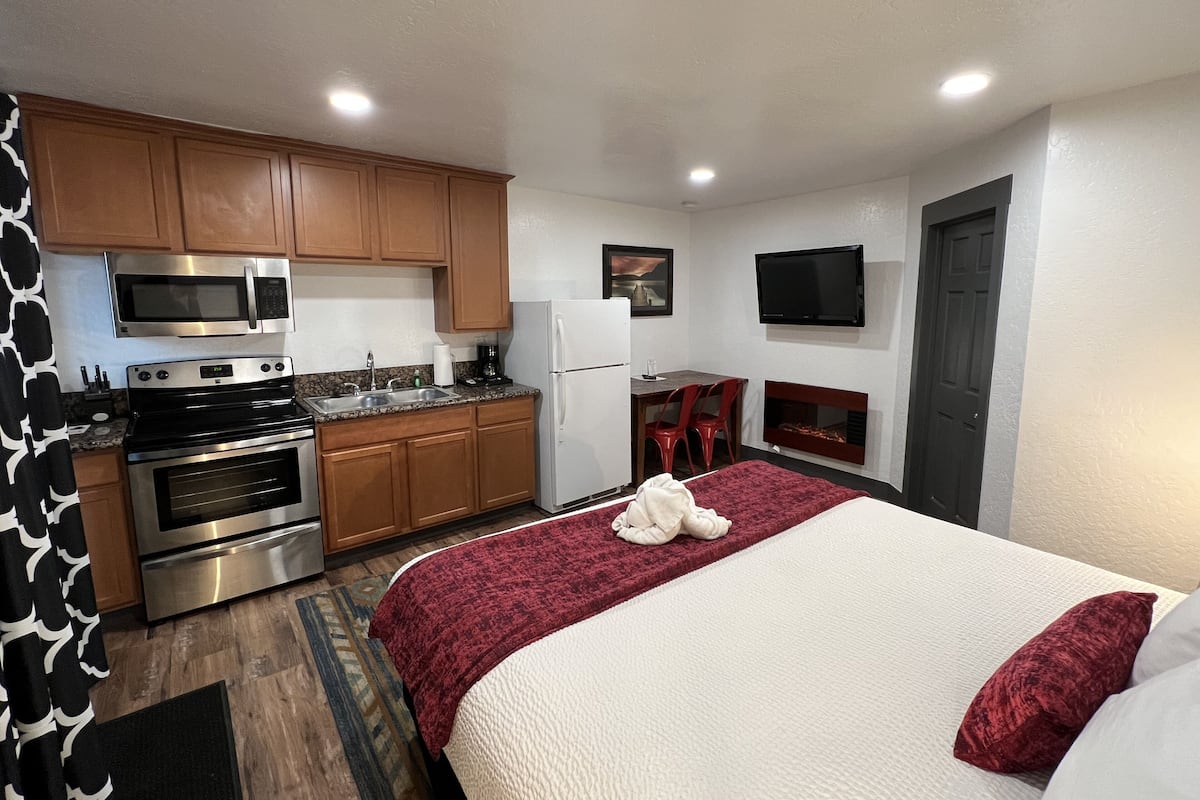 A compact hotel room with a kitchenette, a bed with a red blanket, a TV mounted on the wall, and a fireplace.