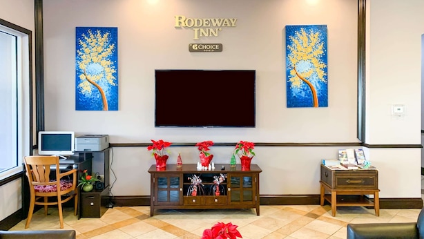A hotel lobby with a television mounted on the wall, a reception desk, and two large paintings of trees. - Rodeway Inn Sun City Center (Sun City Center)