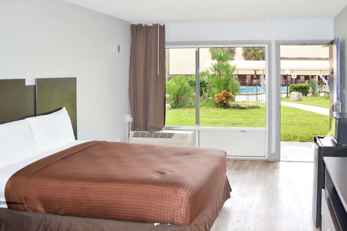 A hotel room with a bed, a desk, and a view of a swimming pool and greenery.