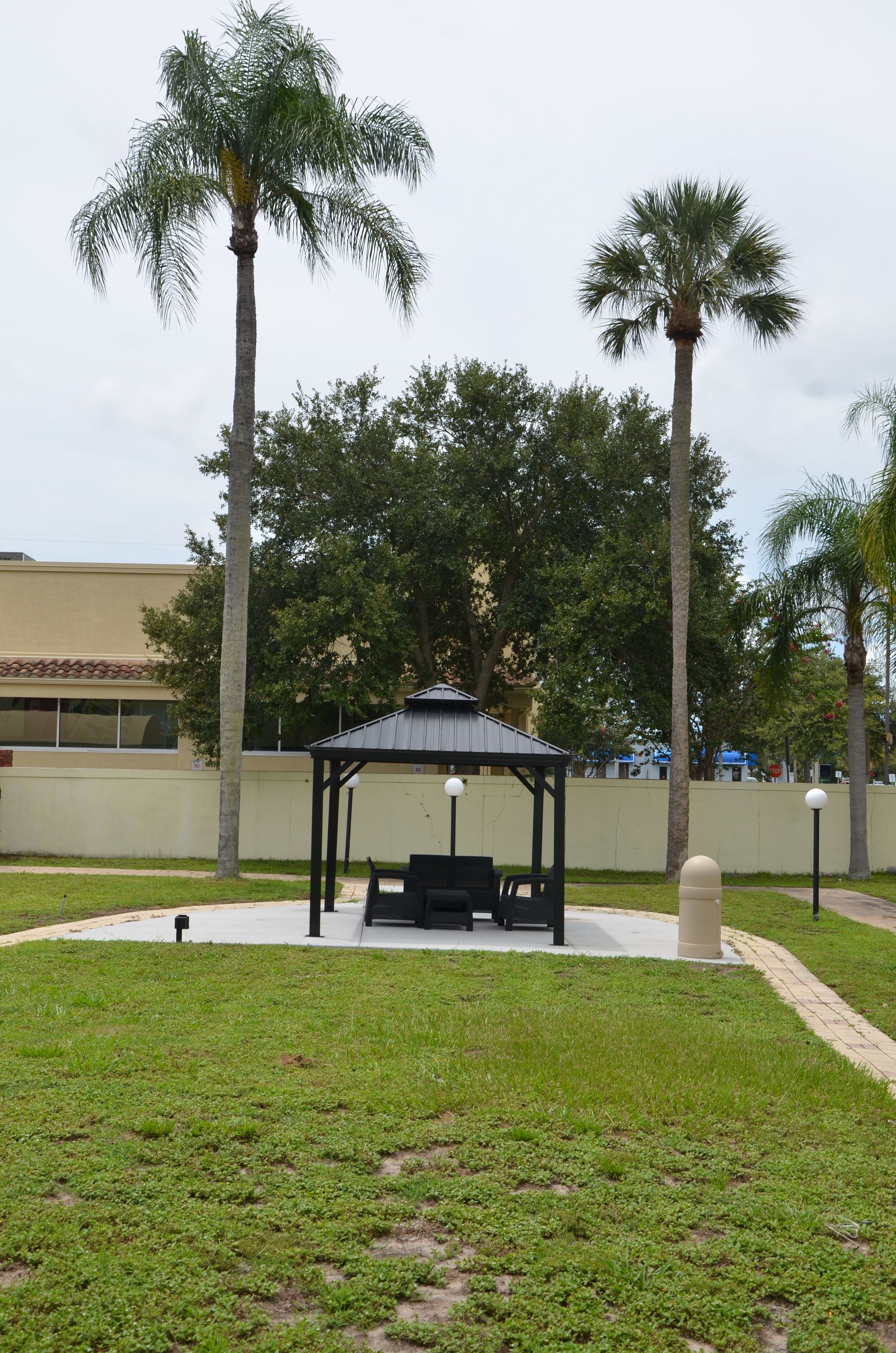 Um gazebo com telhado e assentos, cercado por grama e palmeiras, com um prédio ao fundo.