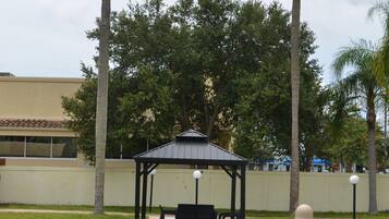 Um gazebo com telhado e assentos, cercado por grama e palmeiras, com um prédio ao fundo.
