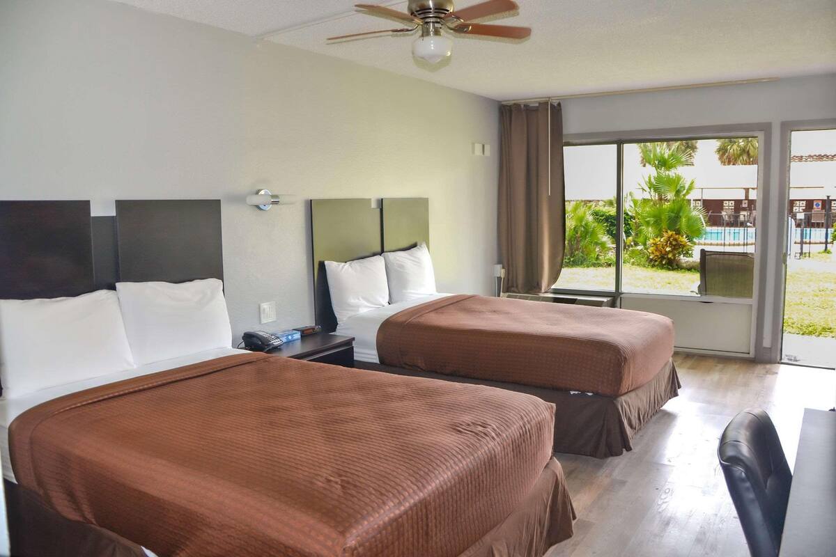 A hotel room with two beds, a ceiling fan, a desk with a chair, and a large window with a view of the outdoors.