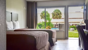 A hotel room with two beds, a desk, and a large window offering a view of palm trees and a building outside. - Rodeway Inn Sun City Center (Sun City Center)